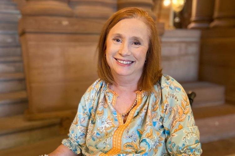 Kimberly Hill, with red hair and a blue-yellow shirt, 坐在轮椅上，坐在奥尔巴尼的国会大厦里, smiling.